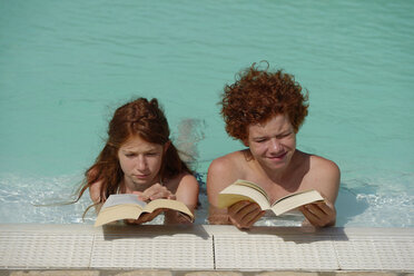 Redheaded girl and boy lying side by side in a swimming pool reading a book - LBF001221