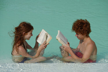 Redheaded girl and boy sitting at pool edge reading a book - LBF001222