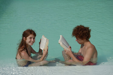 Redheaded girl and boy sitting at pool edge reading a book - LBF001223