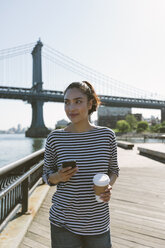 USA, New York City, portrait of smiling young woman with smartphone and coffee to go - GIOF000144