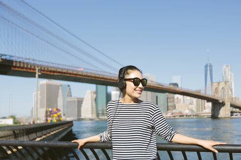 USA, New York City, Porträt einer jungen Frau, die mit Kopfhörern vor der Skyline Musik hört - GIOF000146