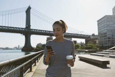 USA, New York City, portrait of smiling young woman with coffee to go looking at her smartphone - GIOF000157