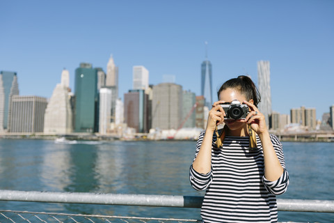 USA, New York City, junge Frau steht vor der Skyline und fotografiert mit der Kamera, lizenzfreies Stockfoto