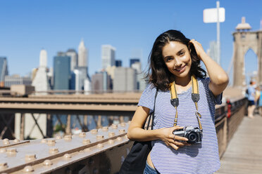 USA, New York City, Porträt einer lächelnden jungen Frau mit Kamera auf der Brooklyn Bridge - GIOF000167