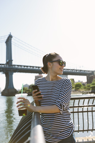 USA, New York City, lächelnde junge Frau mit Softdrink, die auf ihr Smartphone schaut, lizenzfreies Stockfoto