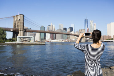 USA, New York City, Rückenansicht einer jungen Frau, die mit ihrem Smartphone die Brooklyn Bridge fotografiert - GIOF000170