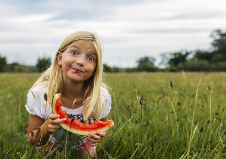Mädchen mit Wassermelonenscheibe auf einer Wiese stehend und ein Gesicht machend - MGOF000800