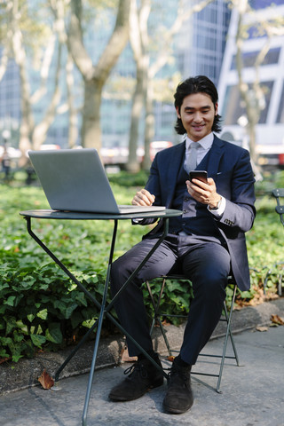 USA, New York City, Manhattan, lächelnder Geschäftsmann mit Laptop und Mobiltelefon im Bryant Park, lizenzfreies Stockfoto