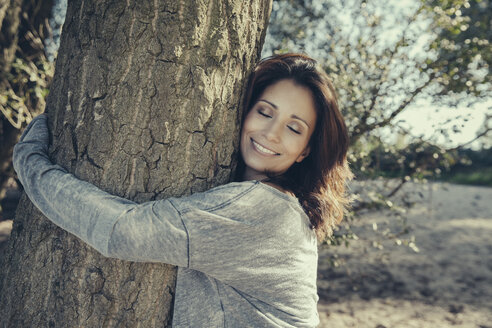Porträt einer lächelnden Frau mit geschlossenen Augen, die einen Baum umarmt - MFF002275