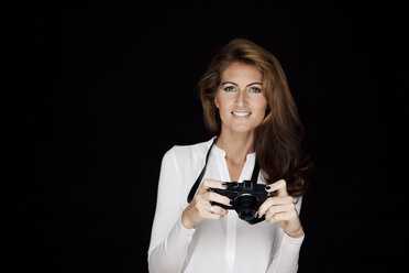 Porträt einer lächelnden Frau mit Kamera vor einem schwarzen Hintergrund - CHAF001542