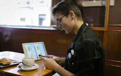 Young woman with digital tablet sitting in a coffee shop looking at her smartphone - MGOF000846