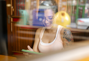 Tätowierte junge Frau sitzt in einem Café und schaut auf ein digitales Tablet - MGOF000855
