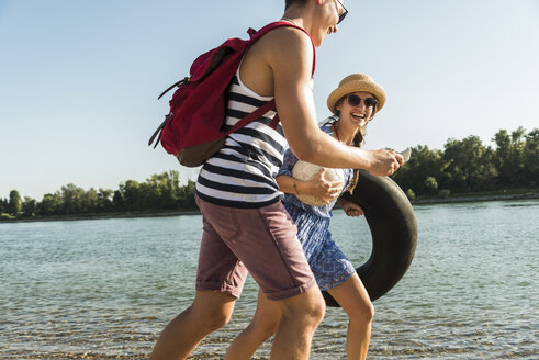 Young couple with inner tube and cell phone walking at the riverside - UUF005850