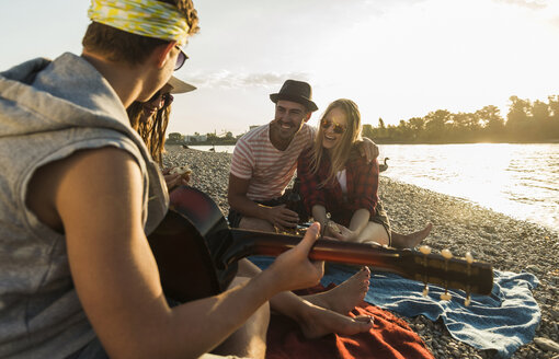Freunde mit Gitarre entspannen sich bei Sonnenuntergang am Flussufer - UUF005905