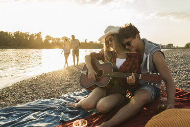Young couple with guitar relaxing at the riverside at sunset - UUF005912
