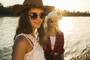Zwei Freunde mit Strohhüten und Sonnenbrillen am Flussufer bei Sonnenuntergang - UUF005921
