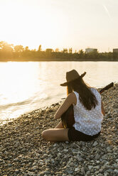 Junge Frau spielt Gitarre am Flussufer bei Sonnenuntergang - UUF005924