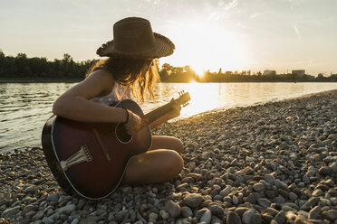 Junge Frau spielt Gitarre am Flussufer bei Sonnenuntergang - UUF005926