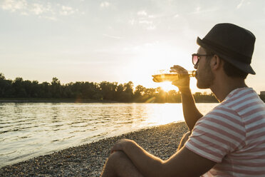 Junger Mann trinkt Bier am Flussufer bei Sonnenuntergang - UUF005927