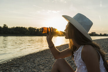 Junge Frau trinkt Bier am Flussufer bei Sonnenuntergang - UUF005929