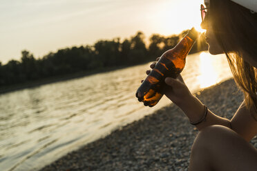 Junge Frau trinkt Bier am Flussufer bei Sonnenuntergang - UUF005930