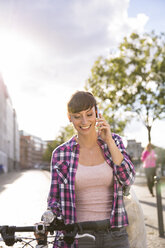 Germany, Berlin, portrait of smiling young woman with bicycle telephoning with smartphone - FKF001403