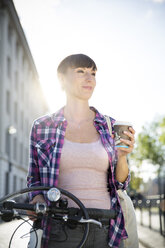 Germany, Berlin, portrait of young woman with bicycle and coffee to go at backlight - FKF001404