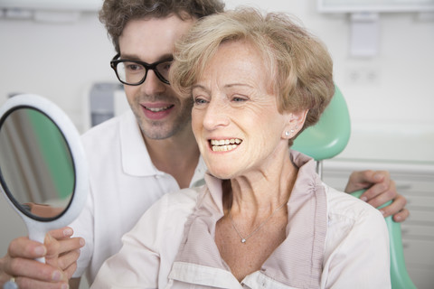 Ältere Frau im Zahnarztstuhl prüft ihre Zähne im Spiegel, lizenzfreies Stockfoto