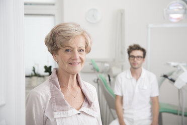 Portrait of smiling senior woman in surgery with dentist in background - FKF001485