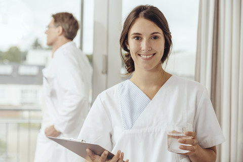 Nurse holding glass of water and clip board, doctor phoning in background stock photo