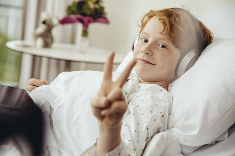 Kranker Junge liegt im Krankenhaus und macht ein Siegeszeichen, trägt Kopfhörer, lizenzfreies Stockfoto