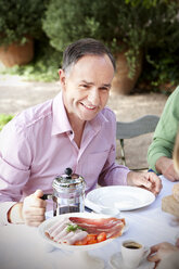 Porträt eines lächelnden Mannes, der am gedeckten Tisch im Garten sitzt und sich mit Freunden unterhält - RMAF000099