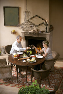 Mature couple sitting at laid table toasting with red wine in front of open fire - RMAF000105