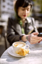 Spanien, Gijon, Tasse Cappucino, junge Frau im Hintergrund mit Smartphone - MGOF000968