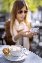 Spain, Gijon, Cup of cappucino, young woman in background using smart phone - MGOF000969