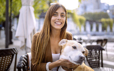 Spain, Gijon, Young woman sitting in pavement cafe, tickling her dog - MGOF000977