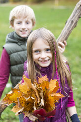 Portrait of girl holding bunch of autumn leaves while her brother standing in the background - SARF002270