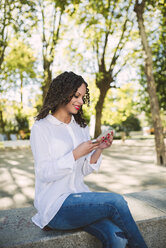 Smiling young woman with brown ringlets and red lips looking at her smartphone - RAEF000611