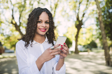 Porträt einer lächelnden jungen Frau mit braunen Locken und roten Lippen mit Smartphone - RAEF000612