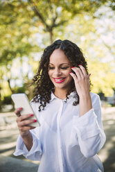 Porträt einer lächelnden jungen Frau mit braunen Locken und roten Lippen, die auf ihr Smartphone schaut - RAEF000613