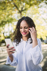 Portrait of smiling young woman with brown ringlets and red lips looking at her smartphone - RAEF000613