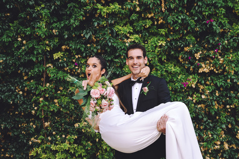 Glücklicher Bräutigam hält aufgeregte Braut in seinen Armen, lizenzfreies Stockfoto
