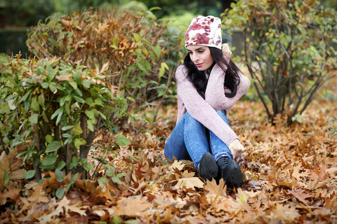 Junge Frau in Herbstmode sitzt auf dem mit Herbstblättern bedeckten Boden, lizenzfreies Stockfoto