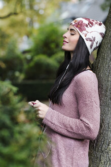 Porträt einer jungen Frau, die sich an einen Baumstamm lehnt und mit Kopfhörern Musik hört - GDF000895