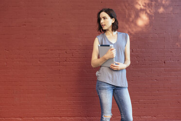Porträt einer Frau mit digitalem Tablet, die vor einer roten Backsteinmauer steht - GIOF000387