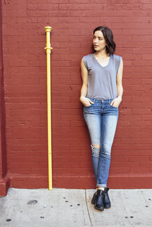 Portrait of woman with hands in her pockets leaning against red brick wall - GIOF000388