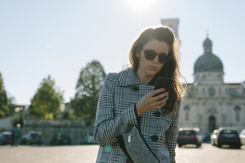 Italien, Vicenza, Frau mit kariertem Mantel und Sonnenbrille schaut auf ihr Handy - GIOF000461