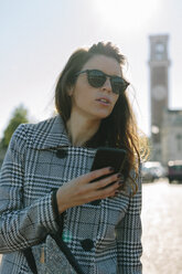 Italy, Vicenza, woman wearing checkered coat and sunglasses holding cell phone - GIOF000462