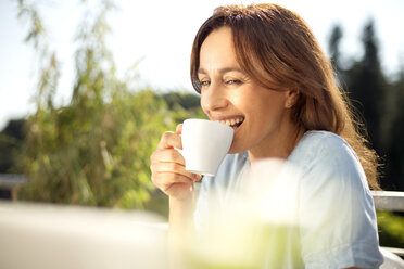 Glückliche Frau auf dem Balkon mit einer Tasse Kaffee und einem Laptop - TOYF001440