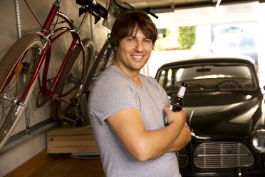 Smiling man in garage with bottle of beer and vintage car - TOYF001467